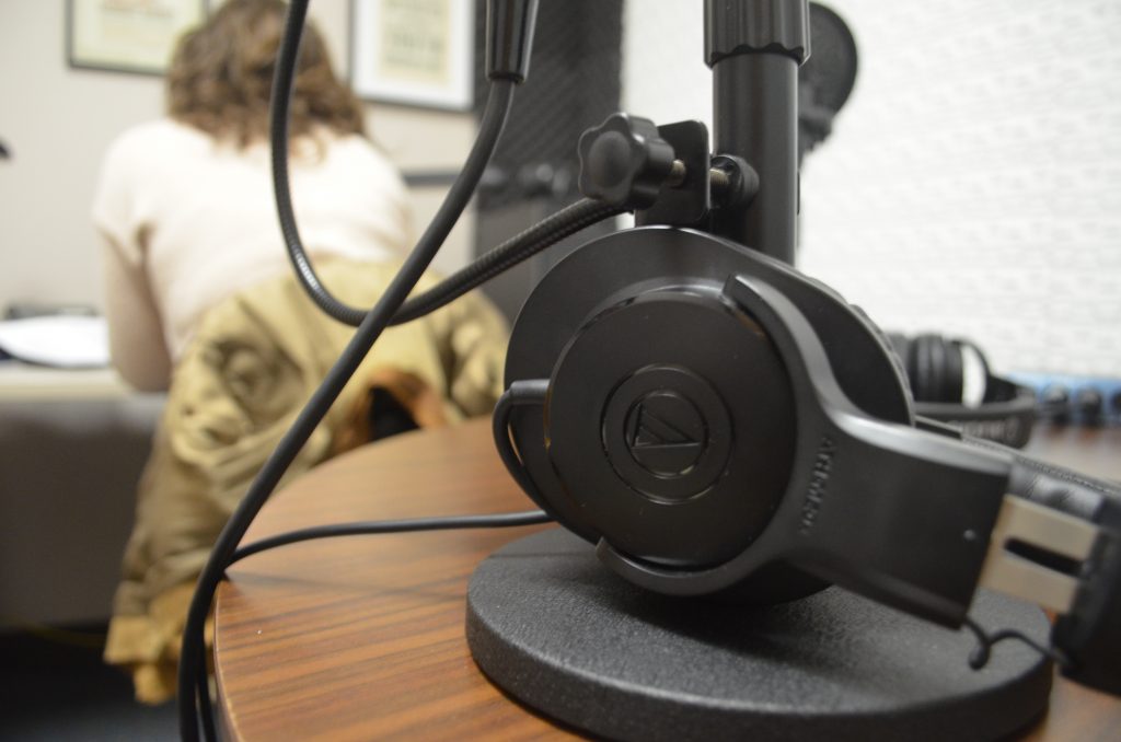 A pair of headphones in the foreground, with a person working at a computer in the background.