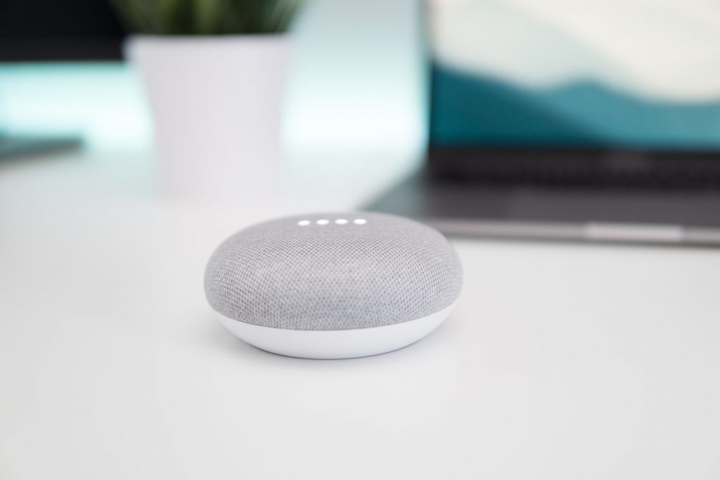 A Google Home Mini smart speaker sitting on a desk in front of a laptop and potted plant.