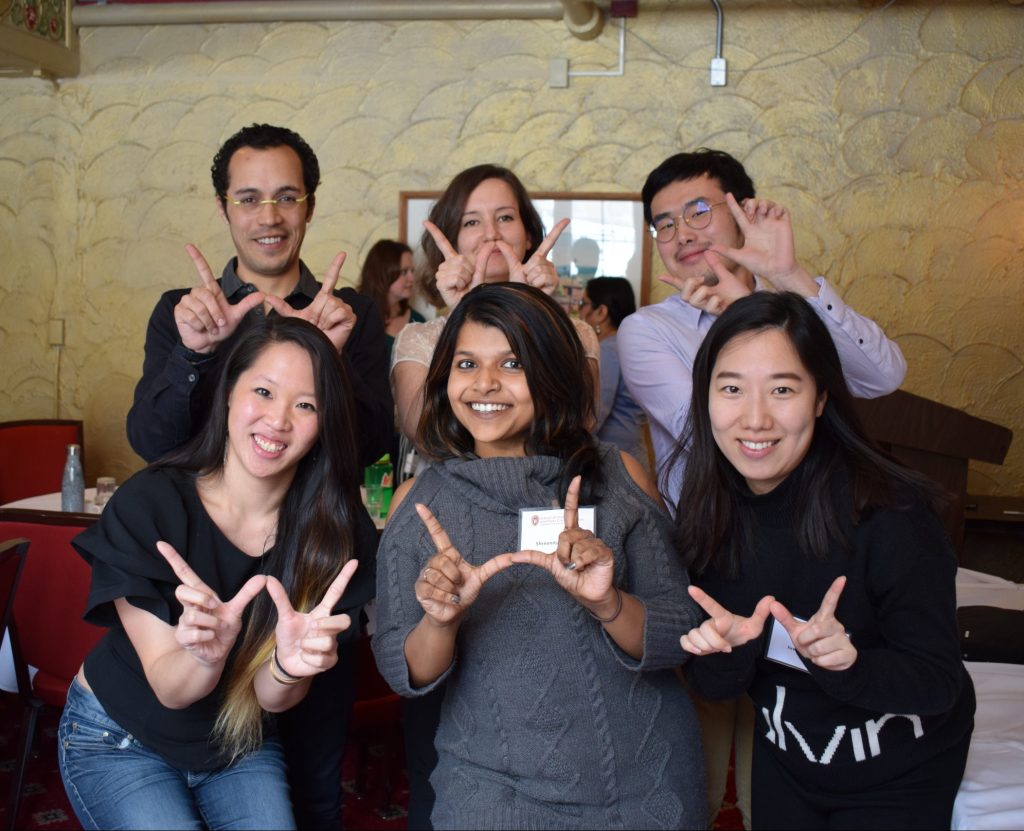 Six SJMC graduate students stand in two rows holding their index fingers and thumbs up in the shape of the Wisconsin W.
