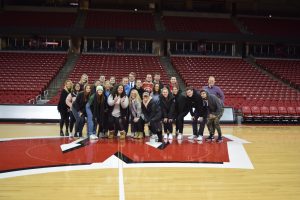 A group of students stands on the center of the basketball court at the Kohl Center on a large motion W logo.
