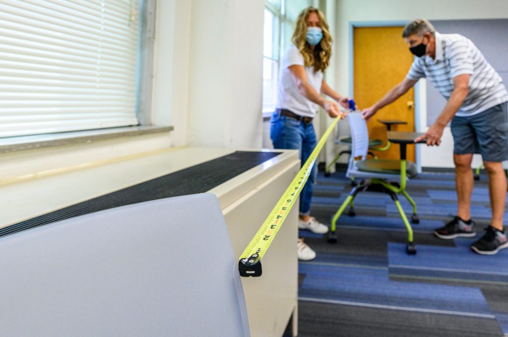 A man and a woman wearing masks measure the distance between two student desks with a tape measure in order to ensure proper distancing.