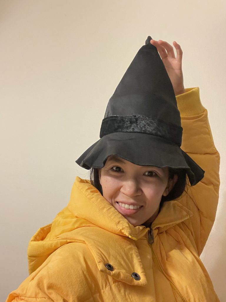 A woman in a yellow coat wearing a witch's hat, holding up the point of the hat over her head.