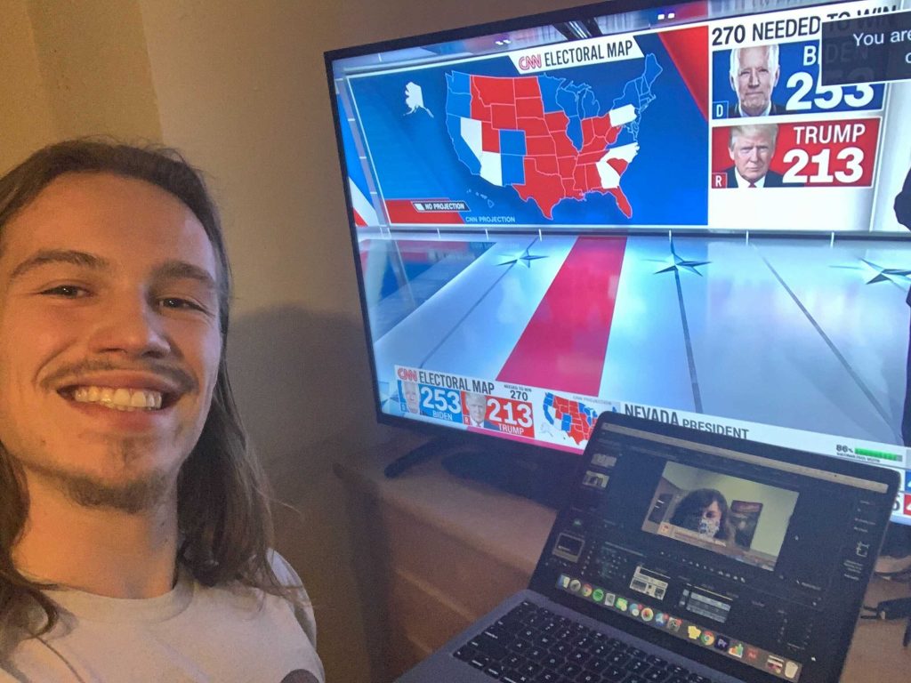 A man facing the camera next to a television screen displaying election results.