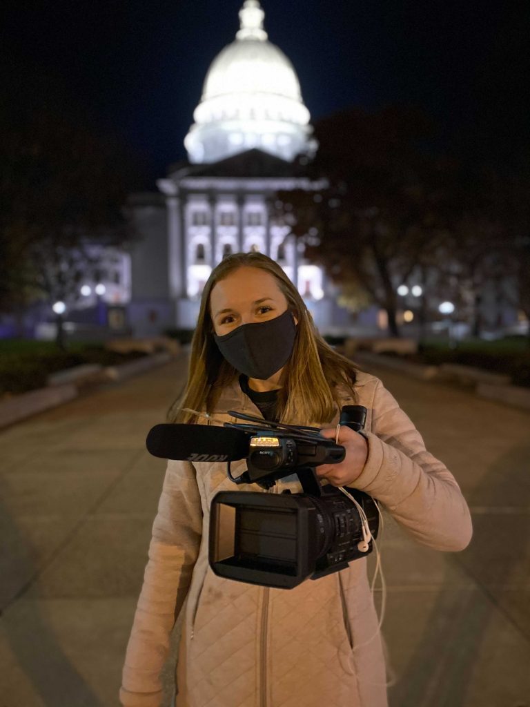 A woman in a face mask holding a video camera stands in front of the Wisconsin Capitol Building lit up at night.