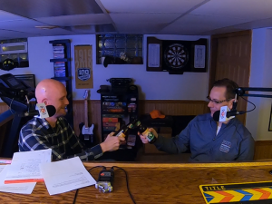 Two men toast with beer bottles sitting behind microphones in a rec room.