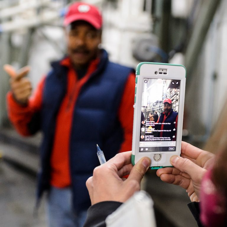 Hands holding a phone being used to live stream a video on Facebook. In the background, Professor Eric Wilcots is out of focus.