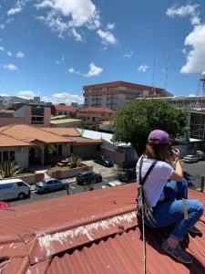 A woman sitting on the roof of a house with her back to the camera. She is holding a camera raised to her eye.