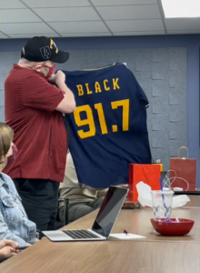 Dave Black holding up a Brewers jersey that reads "Black 91.7"