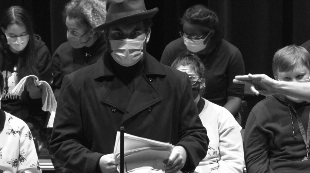 Black and white photo of a man in a mask and fedora holding a script and reading into a mic.