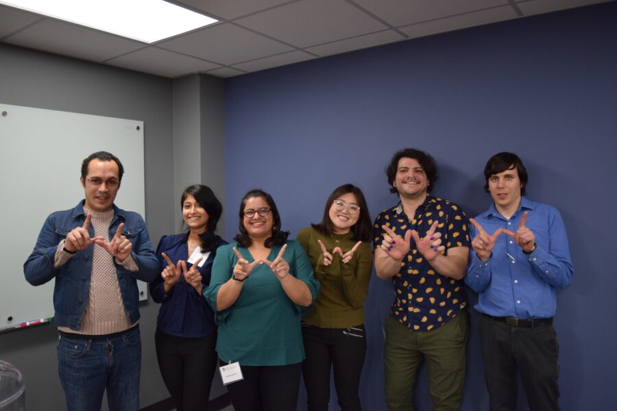 SJMC graduate students stand against a gray wall making a "W" in front of their bodies with their hands.