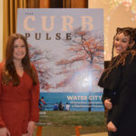 Two female students stand on either side of a foam board showing the cover of Curb magazine's "Pulse" issue at the Curb launch party on December 13, 2022 at the Memorial Union.